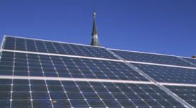 Vista frontal en primer plano de una placa solar sobre un cielo azul donde se entrevé lo alto de una torre de una Iglesia