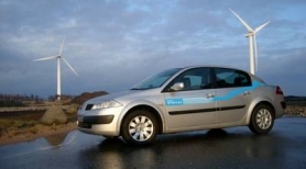 Vehículo eléctrico circulando por una carretera. Al fondo se ven dos aerogeneradores