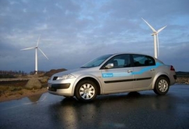 Vehículo eléctrico circulando por una carretera. Al fondo se ven dos aerogeneradores
