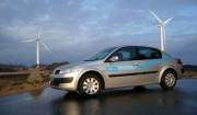 Vehículo eléctrico circulando por una carretera. Al fondo se ven dos aerogeneradores