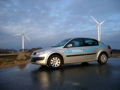 Vehículo eléctrico circulando por una carretera. Al fondo se ven dos aerogeneradores