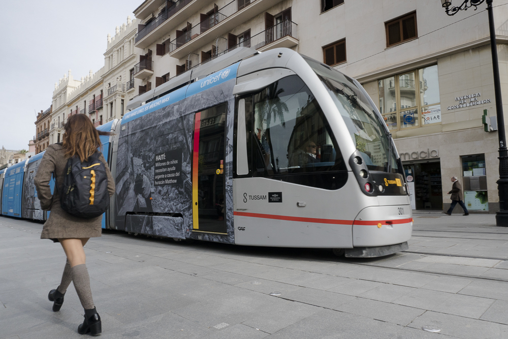 Vista de espalda de una estudiante que va andando por la ciudad al lado de un tranvía 